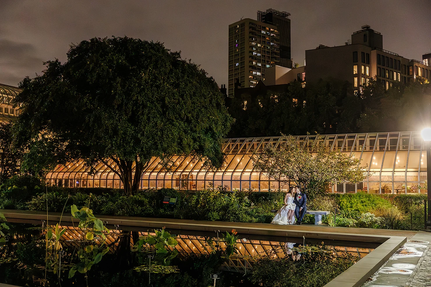 nyc Palm House at the Brooklyn Botanic Garden non traditional wedding dj Bride and groom sit on a bench in a glowing night garden, beside a greenhouse and city high-rises reflected in water