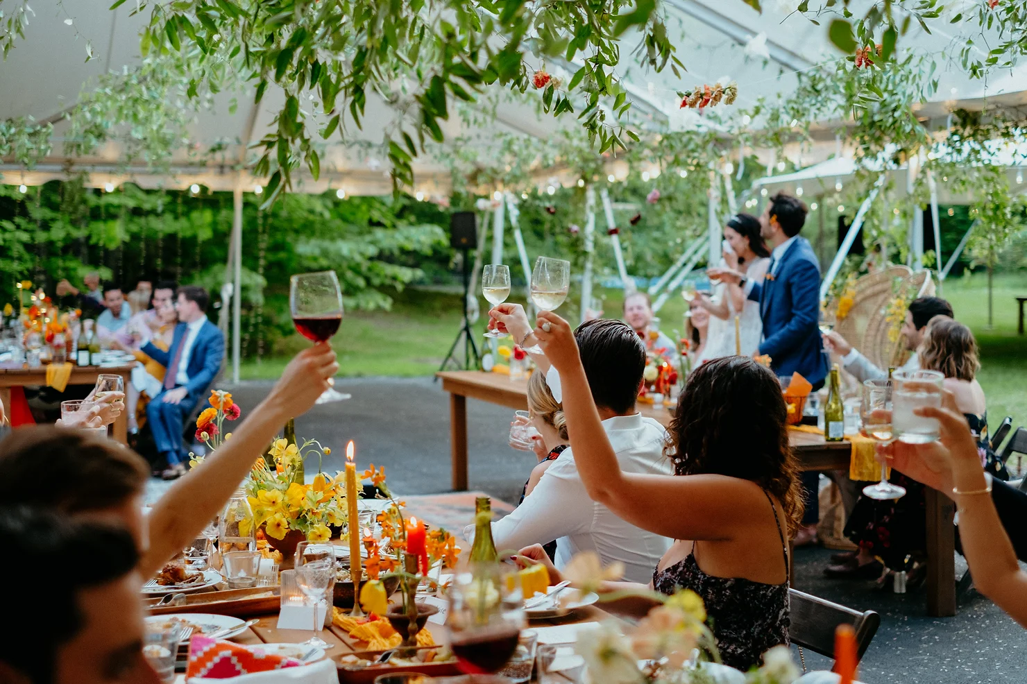 non traditional wedding dj Catskills: Guests raise wine glasses at an outdoor wedding reception under a leafy tent, with floral tables and a smiling couple in blue suits