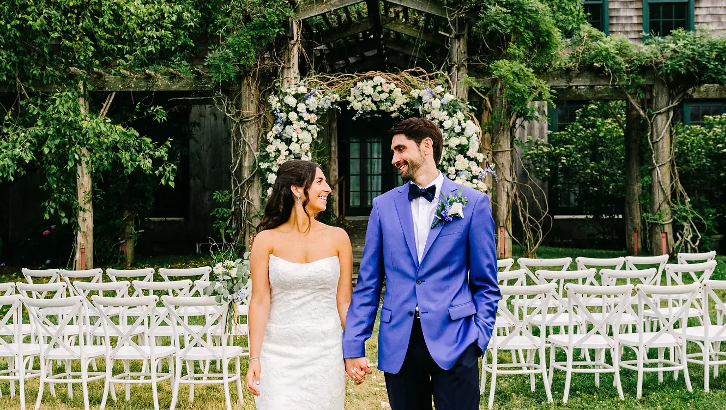 NYC Wedding DJ Bride in white gown and groom in blue suit smile and hold hands before a flower arch with white chairs at an outdoor wedding.