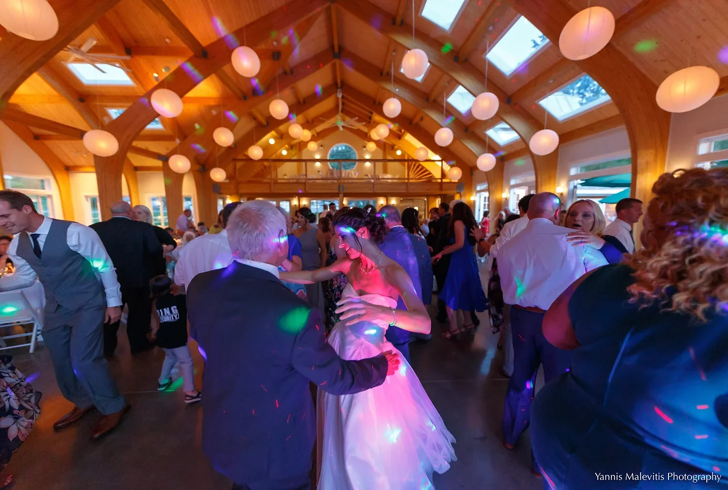 nyc catskills wedding dj Wedding guests dance in a wooden hall under colorful disco lights, with a bride in white and Yannis Malevitis Photography watermark.