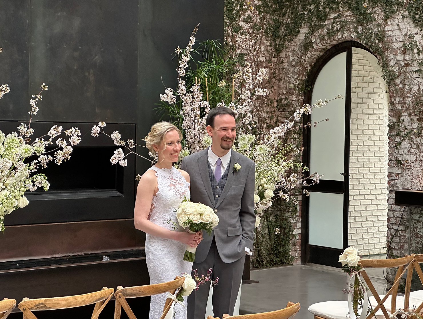 The Foundry nyc non traditional wedding DJ: Bride in white lace dress and groom in gray suit smile with white rose bouquet at an indoor wedding arch of blossoms and ivy