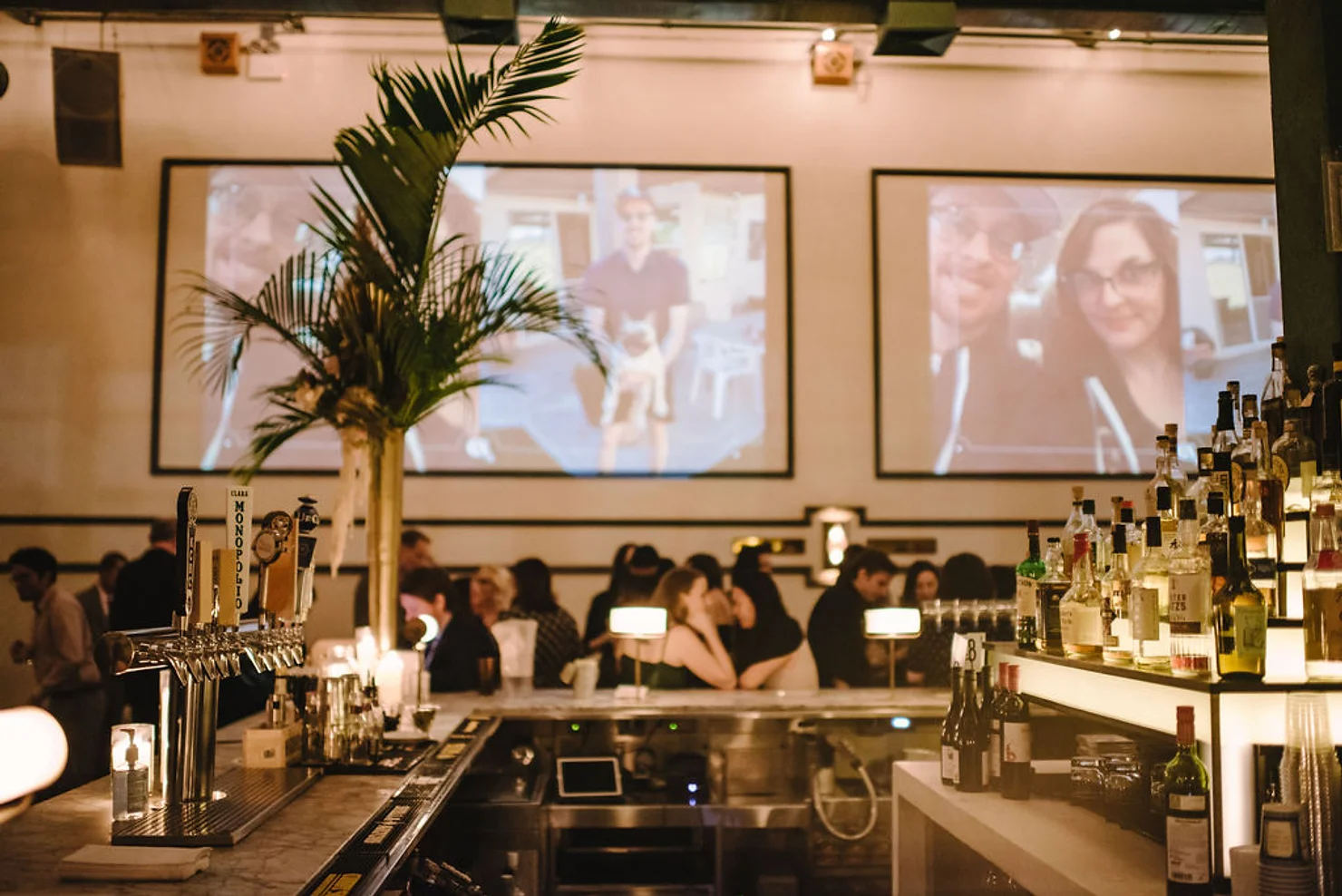 nyc non traditional wedding dj Warm-lit bar with patrons, bottles and taps in foreground, palm tree, and two projected photos; Monopoli tap visible.