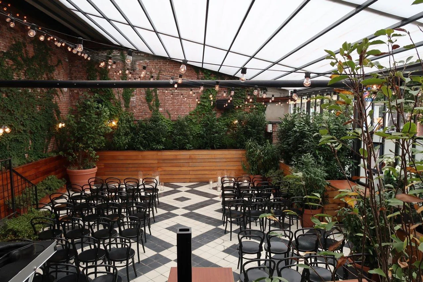 nyc weddind dj wythe hotel Empty greenhouse-style patio with black chairs, string lights, ivy-covered brick walls, and a black-and-white tiled floor