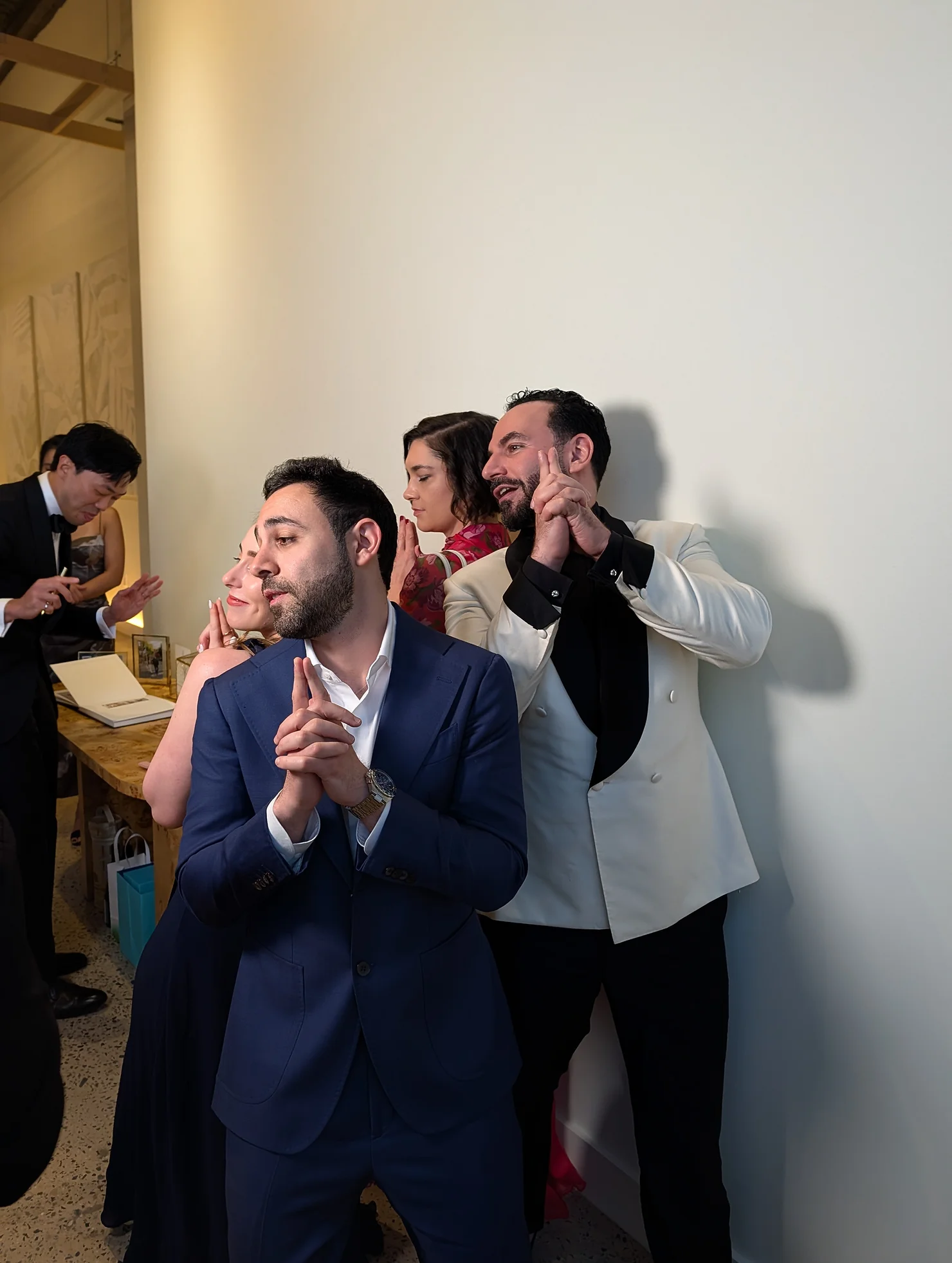 nyc catskills wedding dj People in formal attire pose with hands clasped in a white-walled gallery, including a man in a blue suit and a man in a cream jacket
