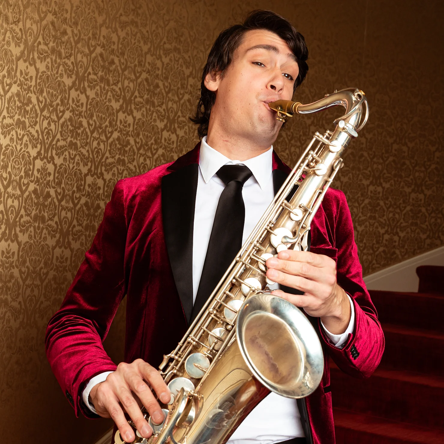 nyc non traditional wedding djs with Live Musicians Man in a burgundy velvet jacket plays a saxophone in front of ornate brown wallpaper, looking focused.
