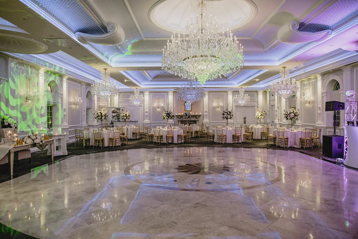 nyc wedding dj bergen county dj Elegant empty ballroom with crystal chandeliers, round tables, gold chairs, floral arrangements, and green-purple lighting.