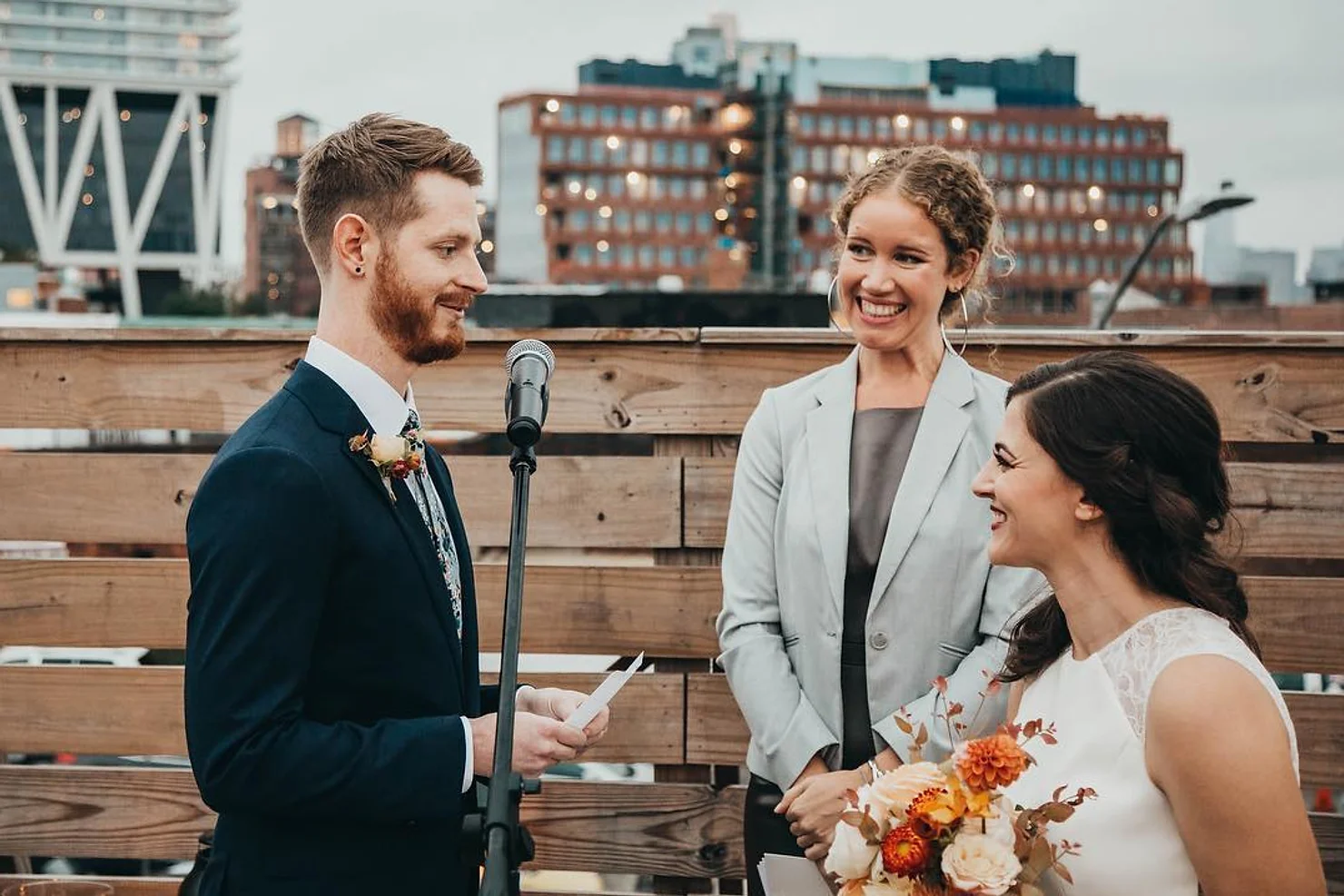 Dobbin St Wedding venue nontraditional wedding dj Smiling couple at rooftop wedding ceremony with officiant and microphone, city buildings in the background, bouquet in view
