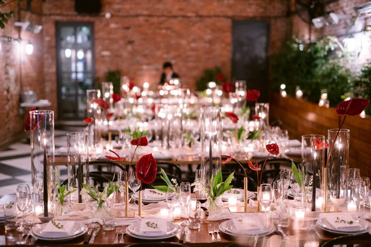 nyc weddind dj wythe hotel Candlelit banquet tables with red flowers and glassware in a brick-walled venue, softly lit and elegant, with a blurred figure in back