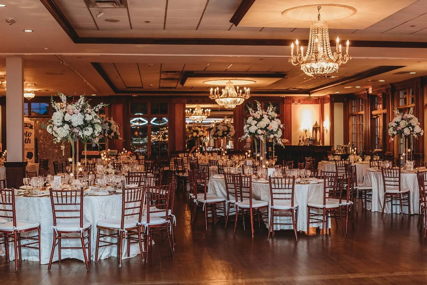 nassau county nyc wedding dj Elegant banquet hall with round set tables, white linens, floral centerpieces, and chandeliers under warm lighting.