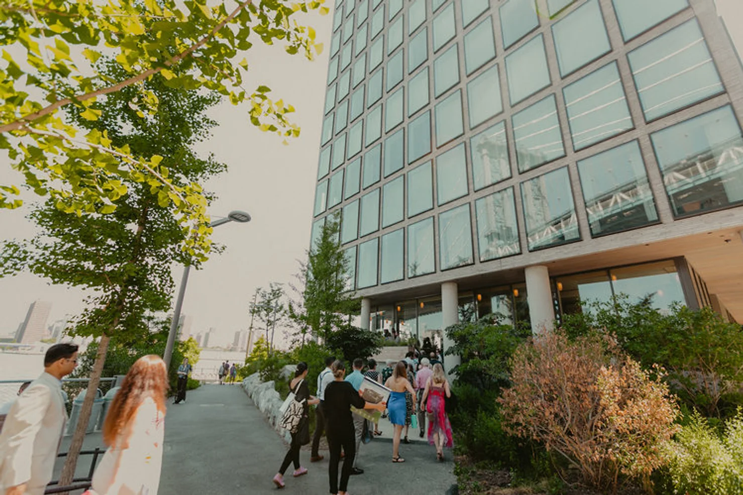 nyc wedding dj People walk toward a modern glass office building lined with trees and shrubs on a bright day, creating a calm urban scene.