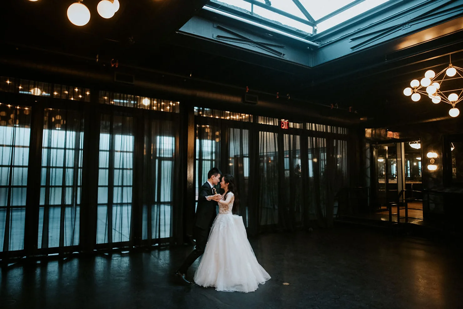 nyc wedding dj Bride and groom dance in a dark modern hall with skylight and chandeliers, while red EXIT signs glow in the background.