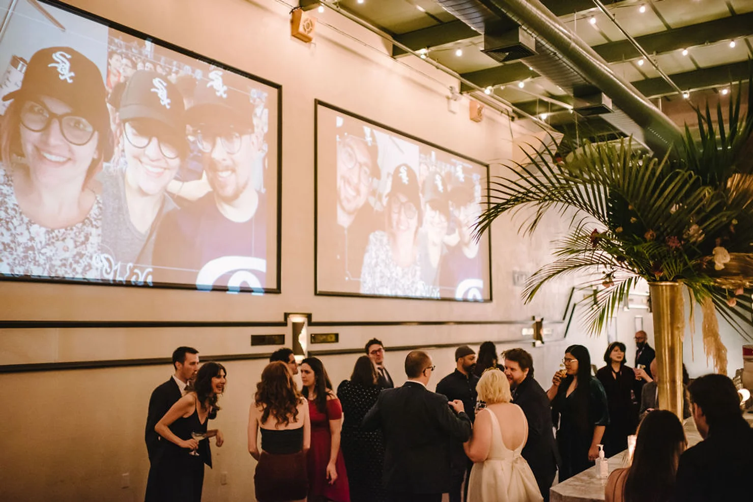 Guests mingle at an indoor event beneath large projected selfies, with warm lighting and a tall palm centerpiece.