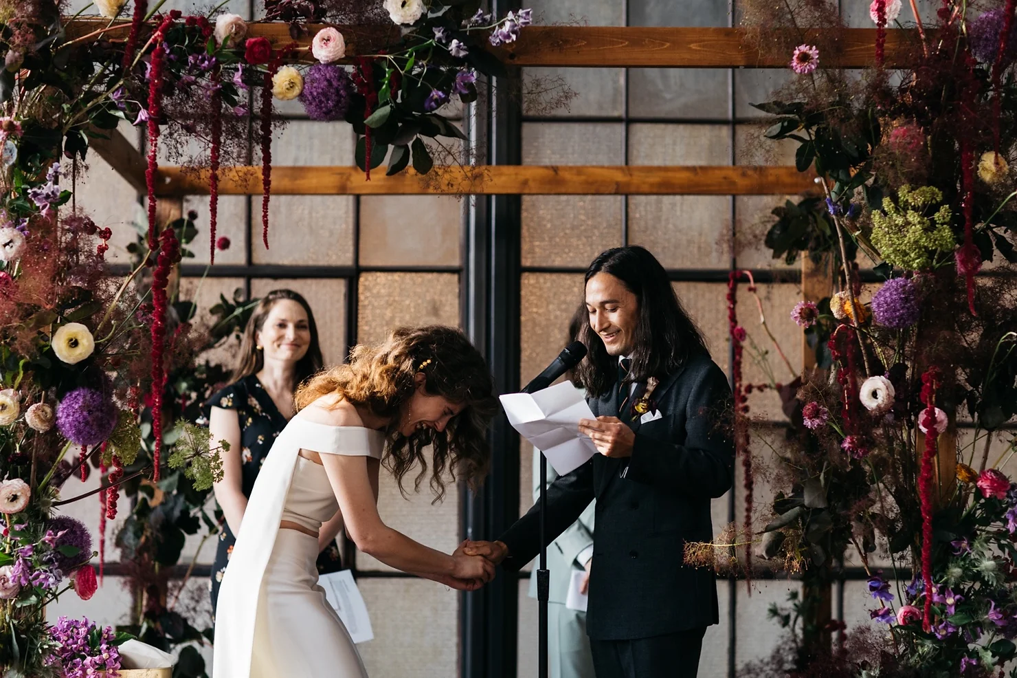 Philly wedding dj Bride and groom hold hands and smile during a wedding ceremony beneath a colorful floral arch while the officiant reads vows