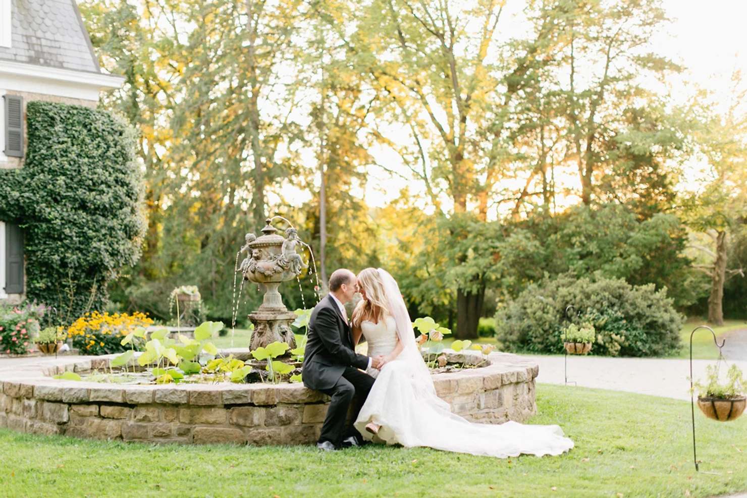 The Inn at Barley Sheaf Farm wedding dj Bride and groom kiss beside a stone fountain in a sunlit garden, with an ivy-covered house and trees in the background.