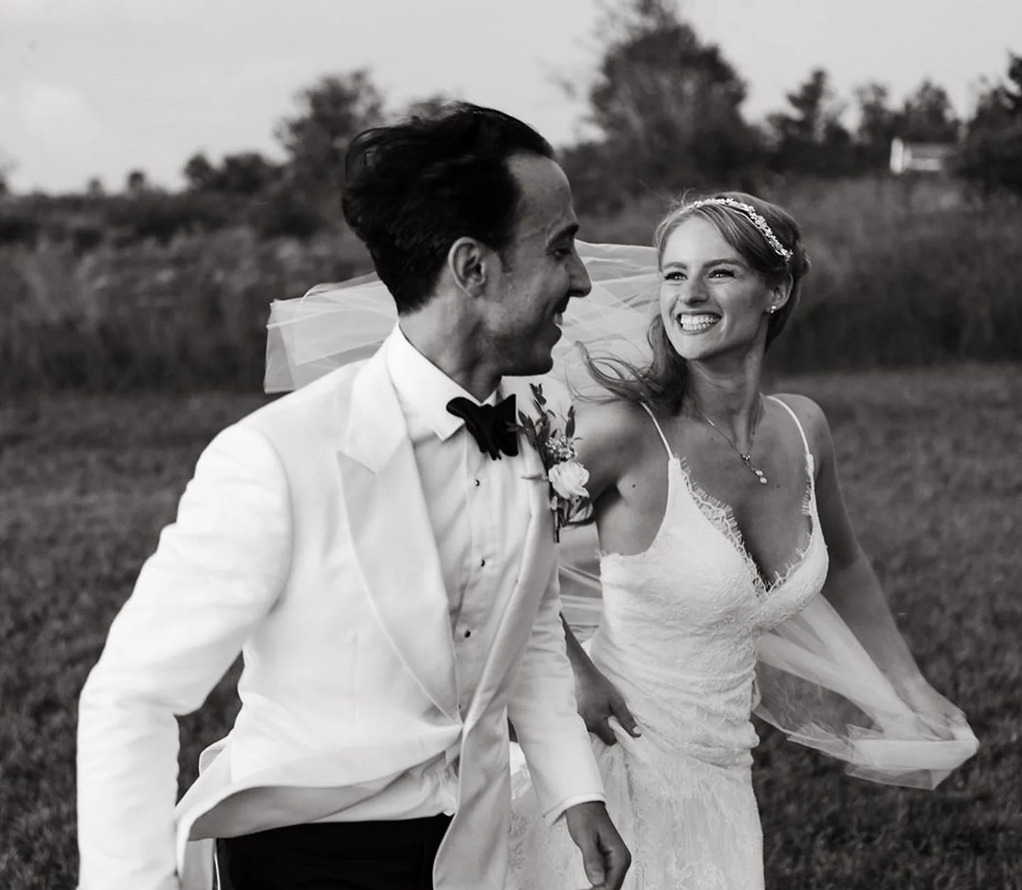 NYC Catskills Wedding DJ Black-and-white wedding portrait of a smiling bride and groom running through a field, bride in lace dress and veil, groom in white tuxedo
