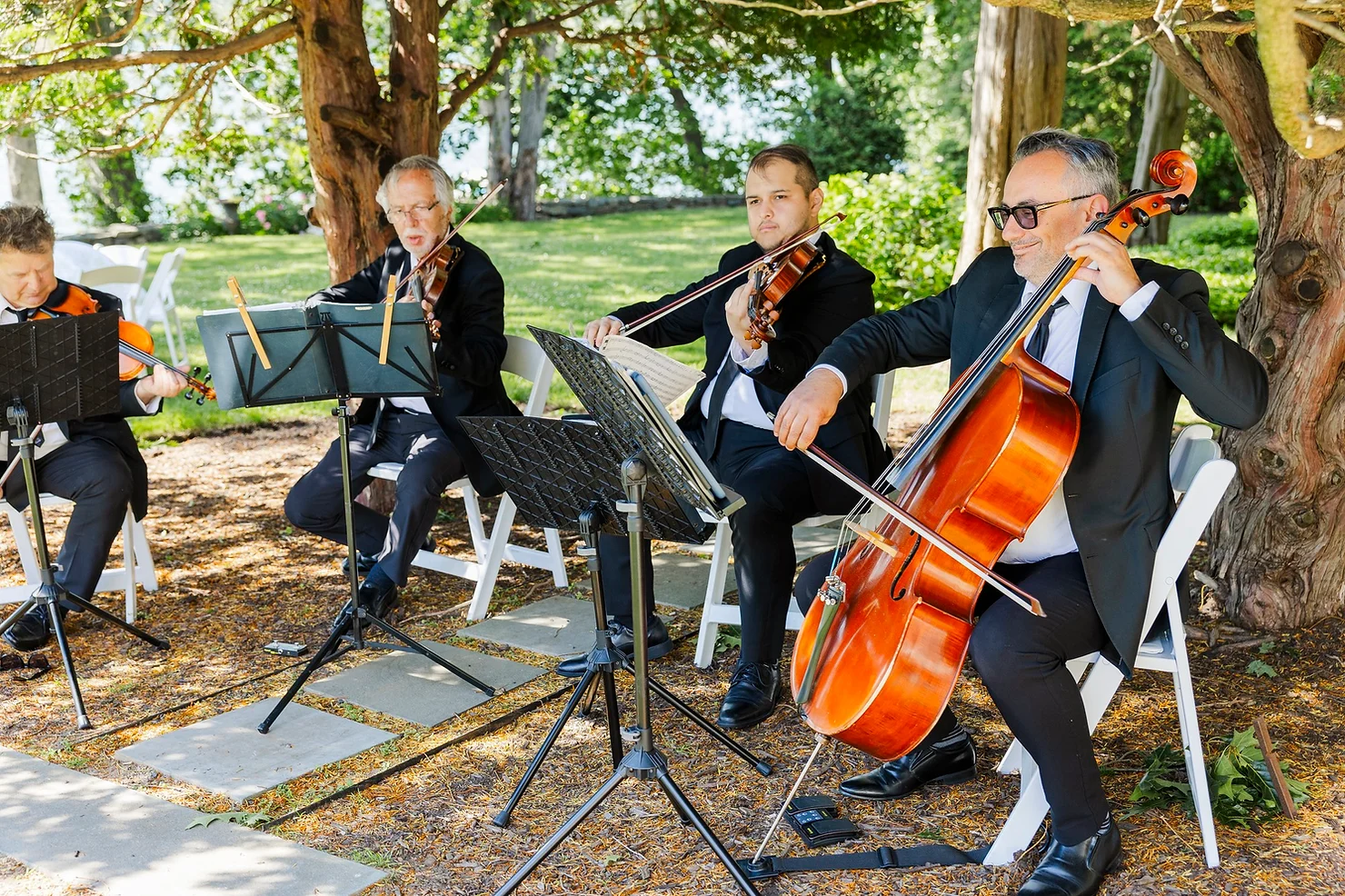 nyc non traditional wedding djs with Live Musicians String quartet in black suits plays outdoors under trees at a garden wedding, with music stands and white chairs nearby.