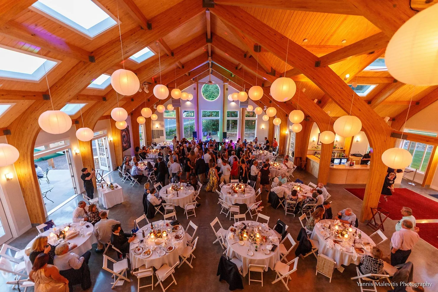 nyc ulster county wedding dj Crowded wedding reception in a wood-beamed hall with hanging globe lights, round tables, guests mingling, and warm festive glow