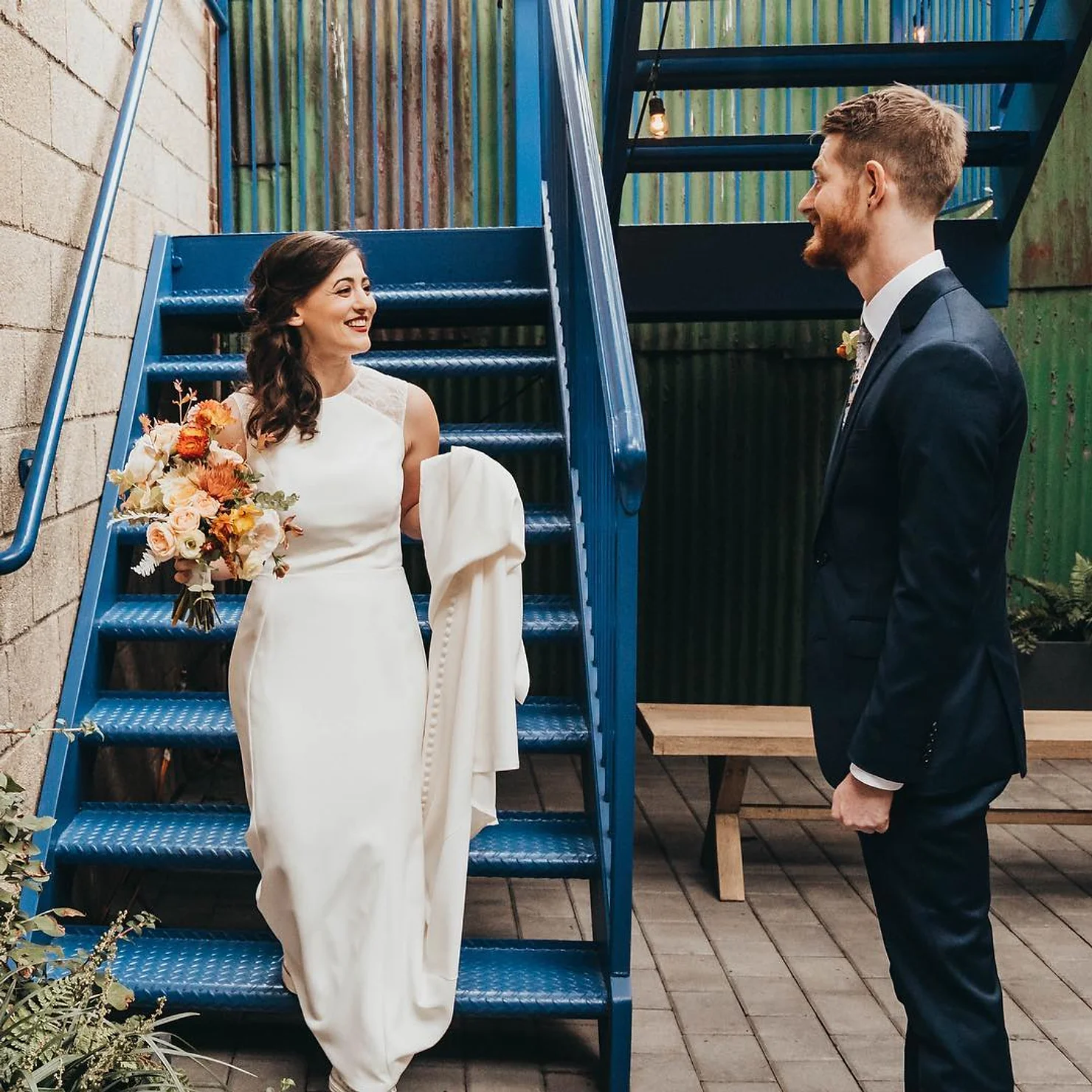Dobbin St Wedding venue non traditional wedding DJ NYC Bride in white dress with bouquet smiles at groom in dark suit on blue metal stairs in an industrial courtyard