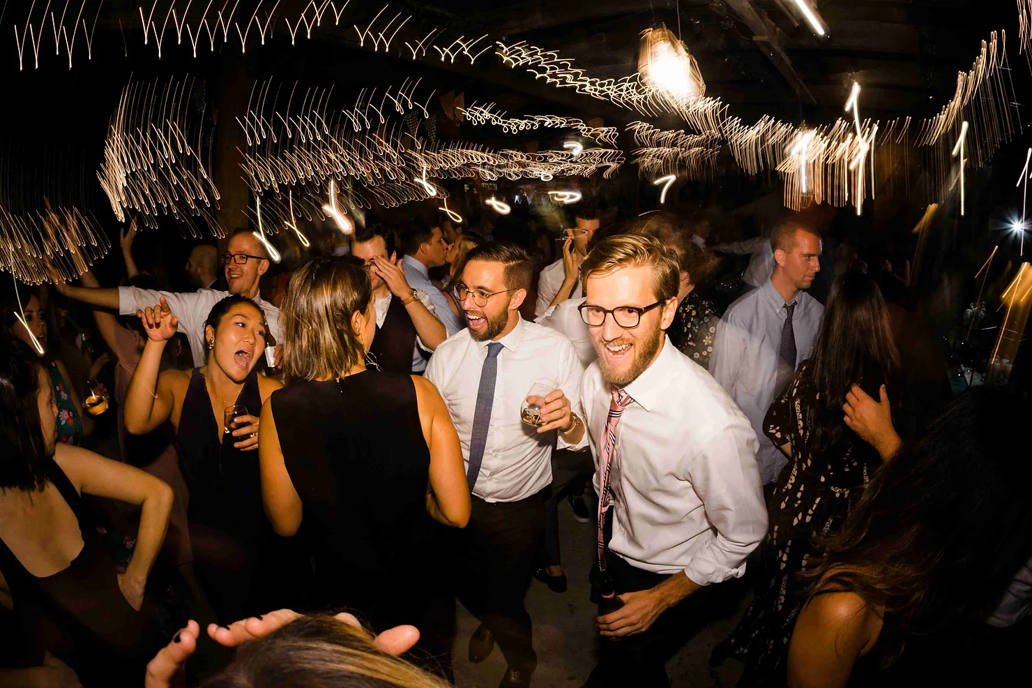 NYC Catskills Wedding DJ Crowd dancing at a lively indoor party under warm string lights, with smiling people in dress shirts and ties holding drinks.