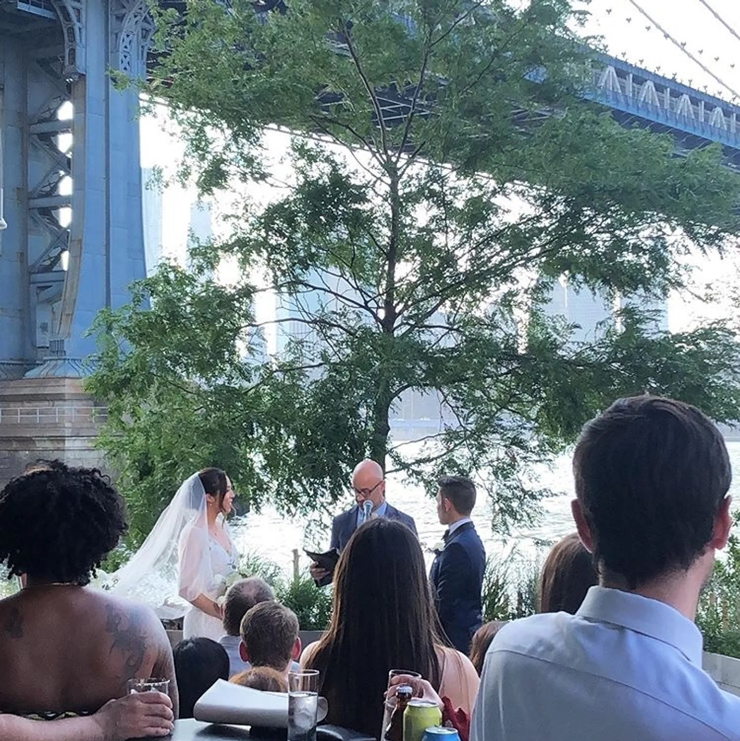 nyc wedding dj Outdoor wedding ceremony by the river under a bridge, bride and groom with officiant before seated guests.