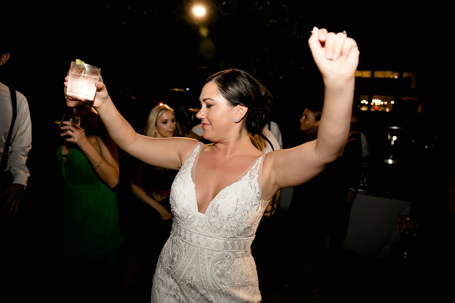 nyc wedding dj the william vale Bride in a white lace dress raises a cocktail at a lively night party, smiling as guests dance in the dark background.