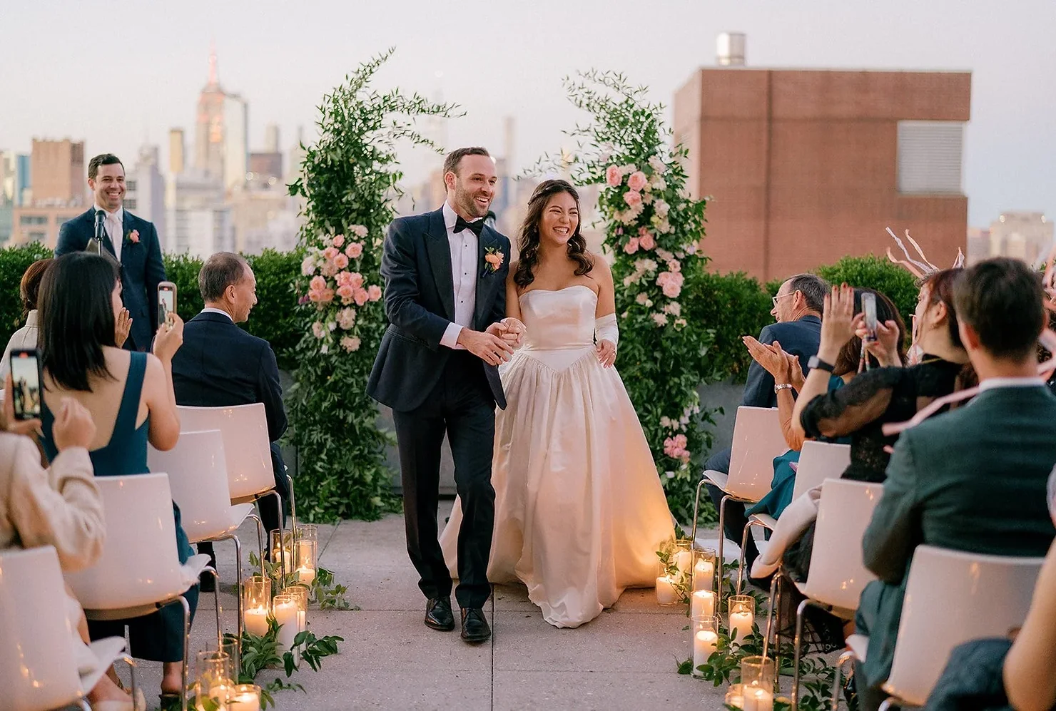 PUBLIC Hotel NYC wedding DJ Rooftop wedding couple walks down aisle, smiling, past floral arches and candles as guests applaud, city skyline behind.