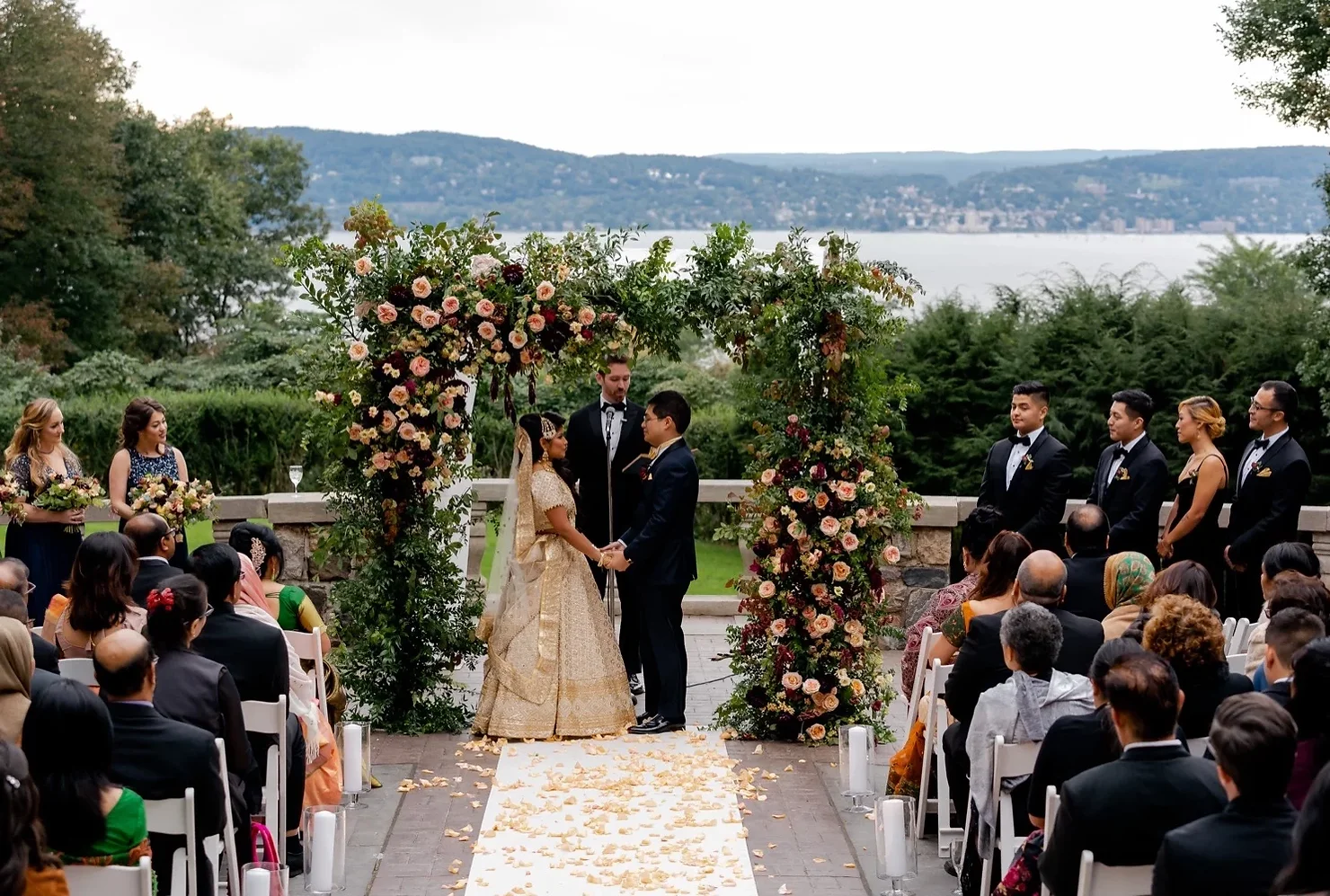 nyc wedding dj Couple standing under a floral arch at an outdoor wedding ceremony on the Tappan Hill Mansion terrace overlooking the Hudson River, guests seated along a petal lined aisle