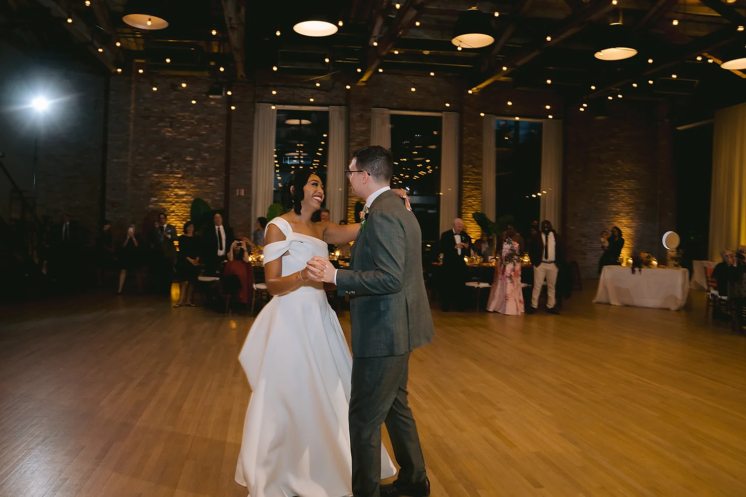 nyc wedding dj Bride in white gown and groom in gray suit slow dance, smiling in a warmly lit reception hall with guests watching.