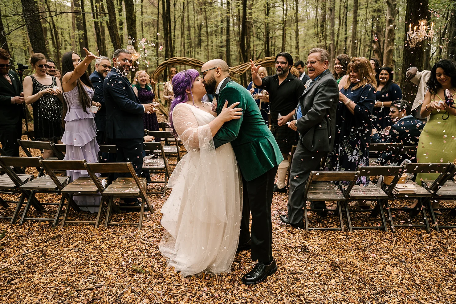 Foxfire Mountain House Wedding DJ: Bride with purple hair and groom in green suit kiss at outdoor woodland wedding as guests cheer and throw confetti