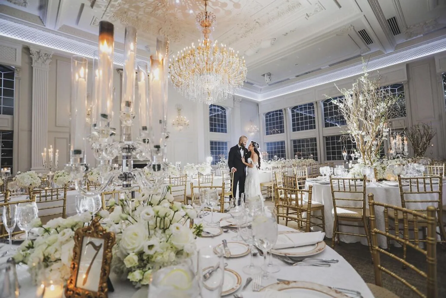 nyc wedding dj bergen county dj Bride and groom embrace in a grand white ballroom with crystal chandeliers, candlelit tables, white flowers, and gold chairs.