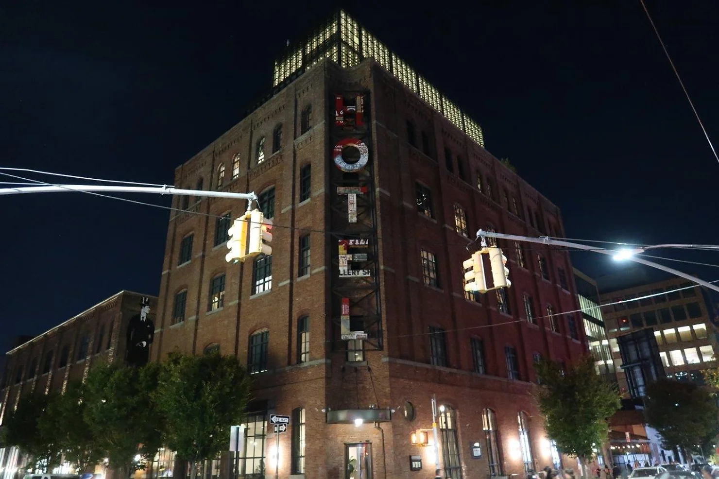 nyc weddind dj wythe hotel Night view of a tall brick building with lit windows and hanging traffic lights on a city street.