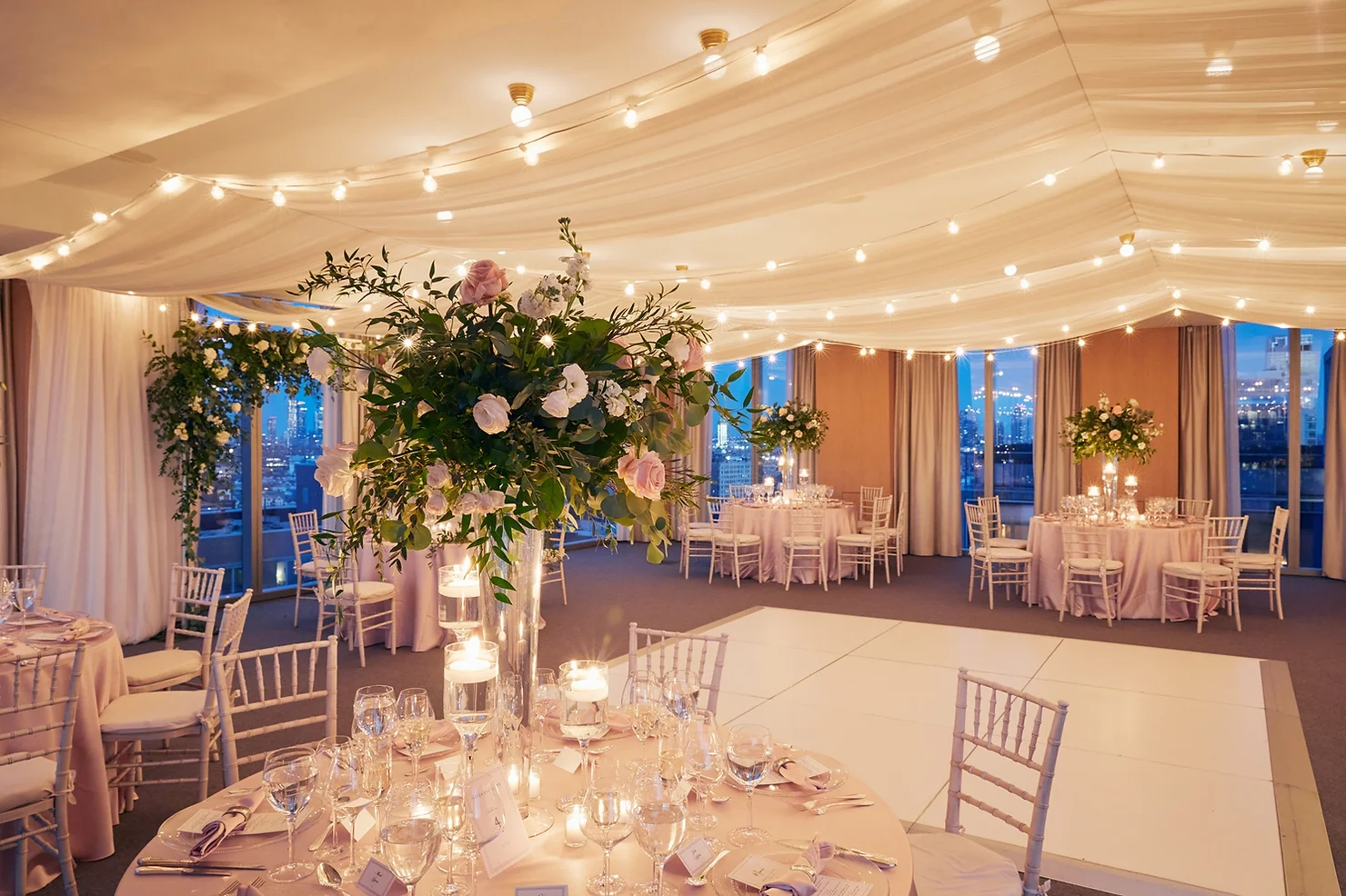 PUBLIC Hotel NYC wedding DJ Elegant wedding reception room with draped string lights, pink floral centerpieces, set tables, and a white dance floor.