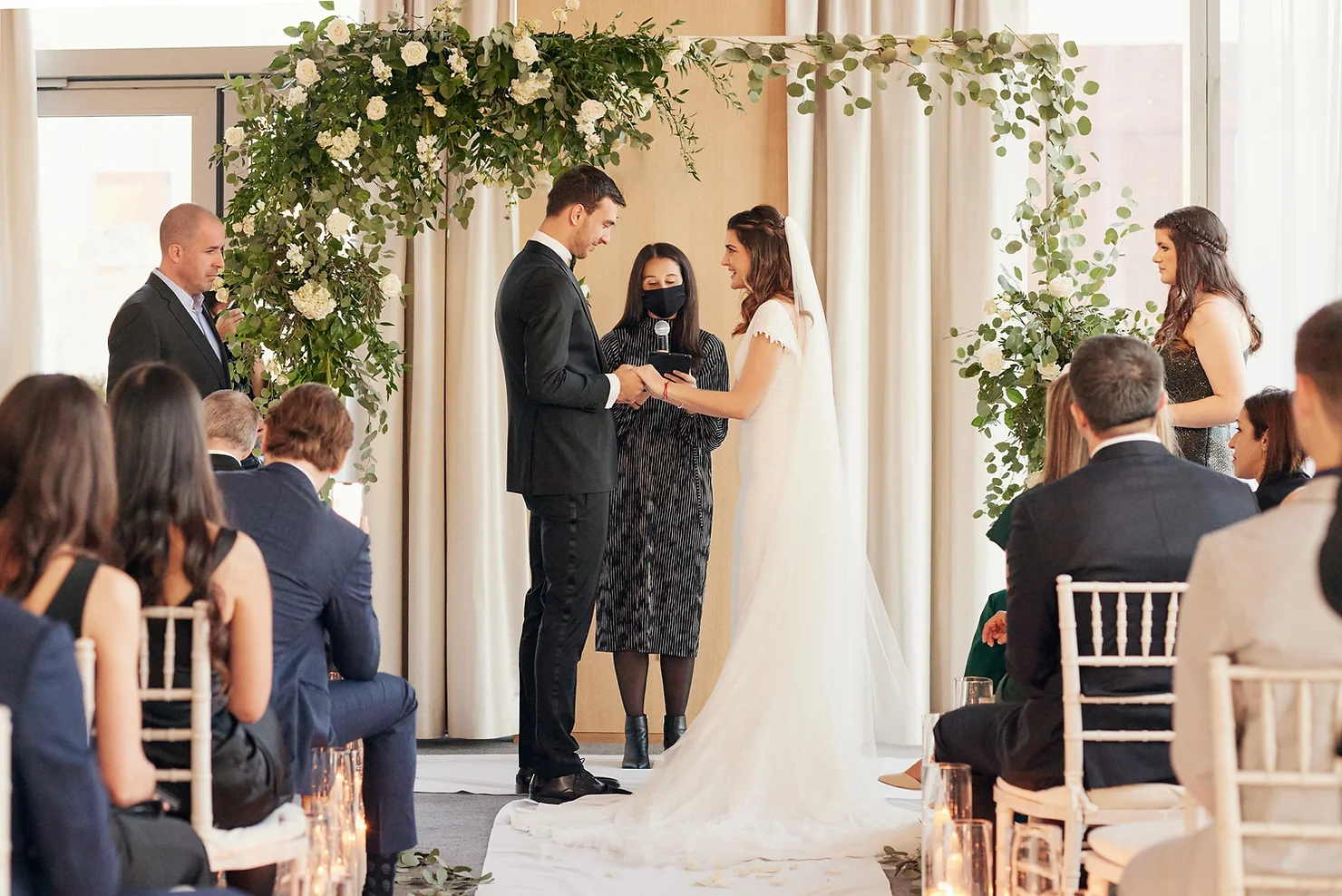PUBLIC Hotel NYC wedding DJ Bride and groom exchange vows under a floral arch as an officiant speaks, with seated guests watching in a bright wedding venue.
