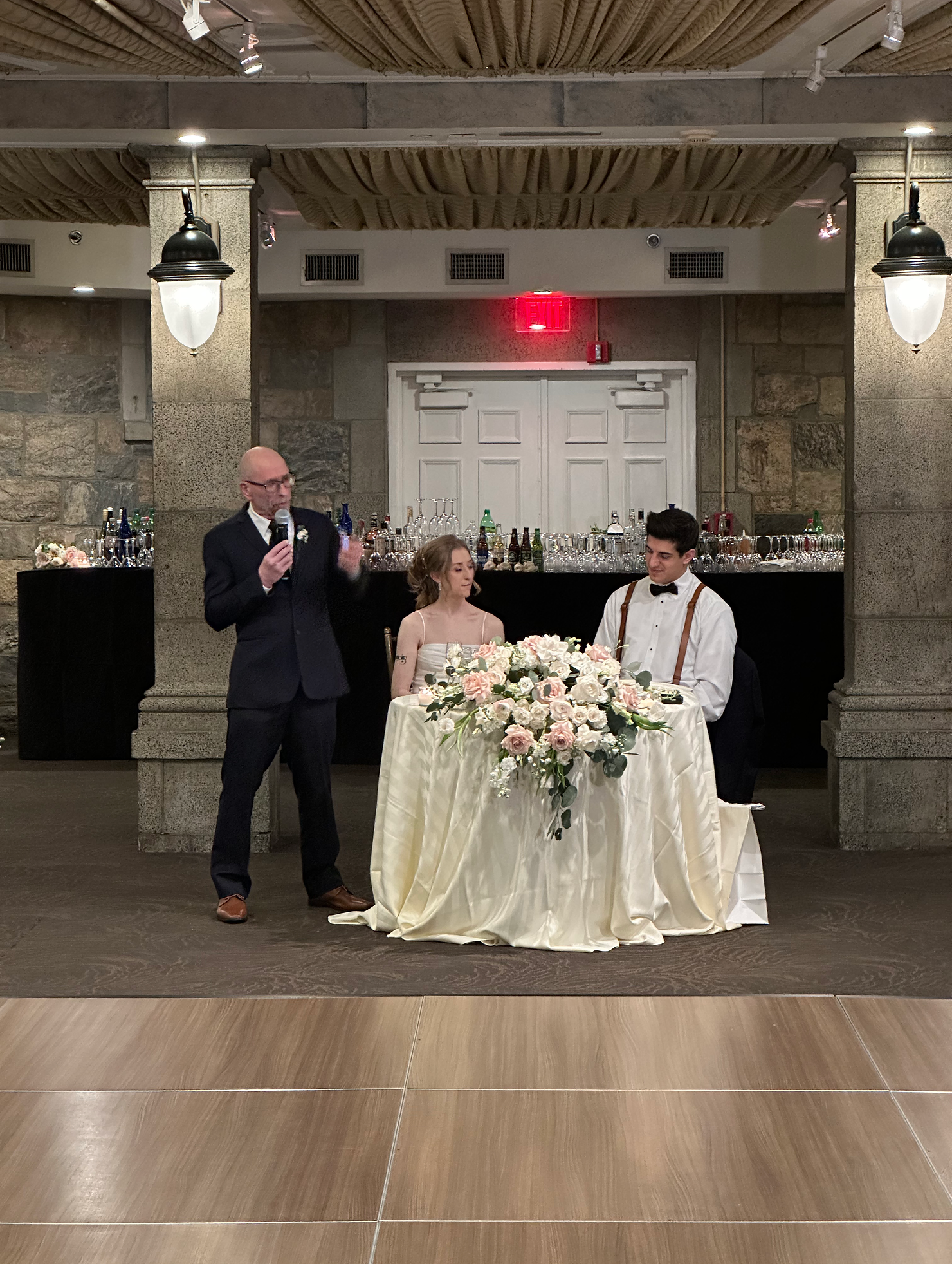 nyc wedding dj Guest giving a wedding toast beside the seated couple at a floral head table inside a Tappan Hill Mansion ballroom in Westchester