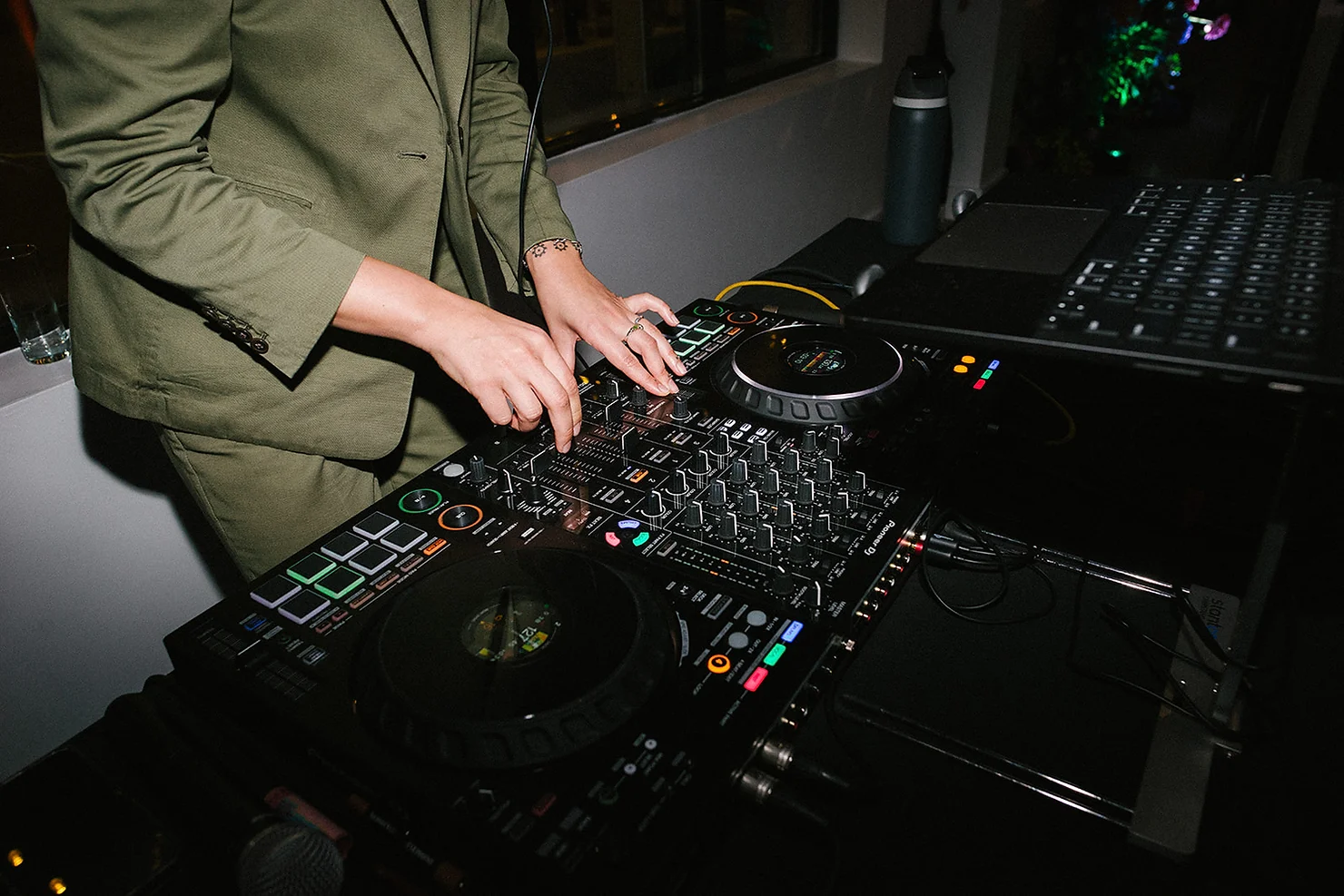 NYC Wedding DJ in a green suit mixes music on a turntable with colorful lights in a dimly lit room, conveying a lively party atmosphere.