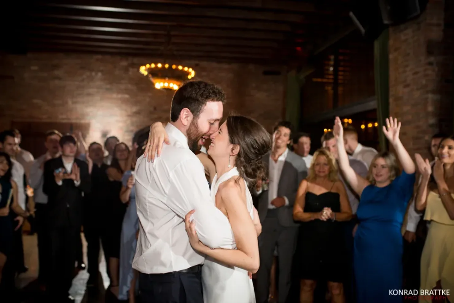nyc wedding dj Wedding couple embraces on a dance floor as guests cheer in a dim brick hall; KONRAD BRATTKE PHOTOGRAPHY watermark.