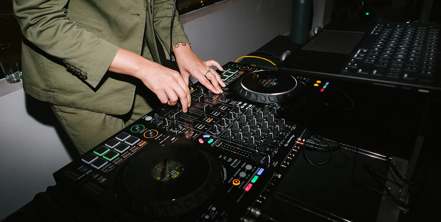 NYC Wedding DJ's hands mixing music on a professional Pioneer DJ controller and laptop during a reception, wearing a tailored olive green blazer.