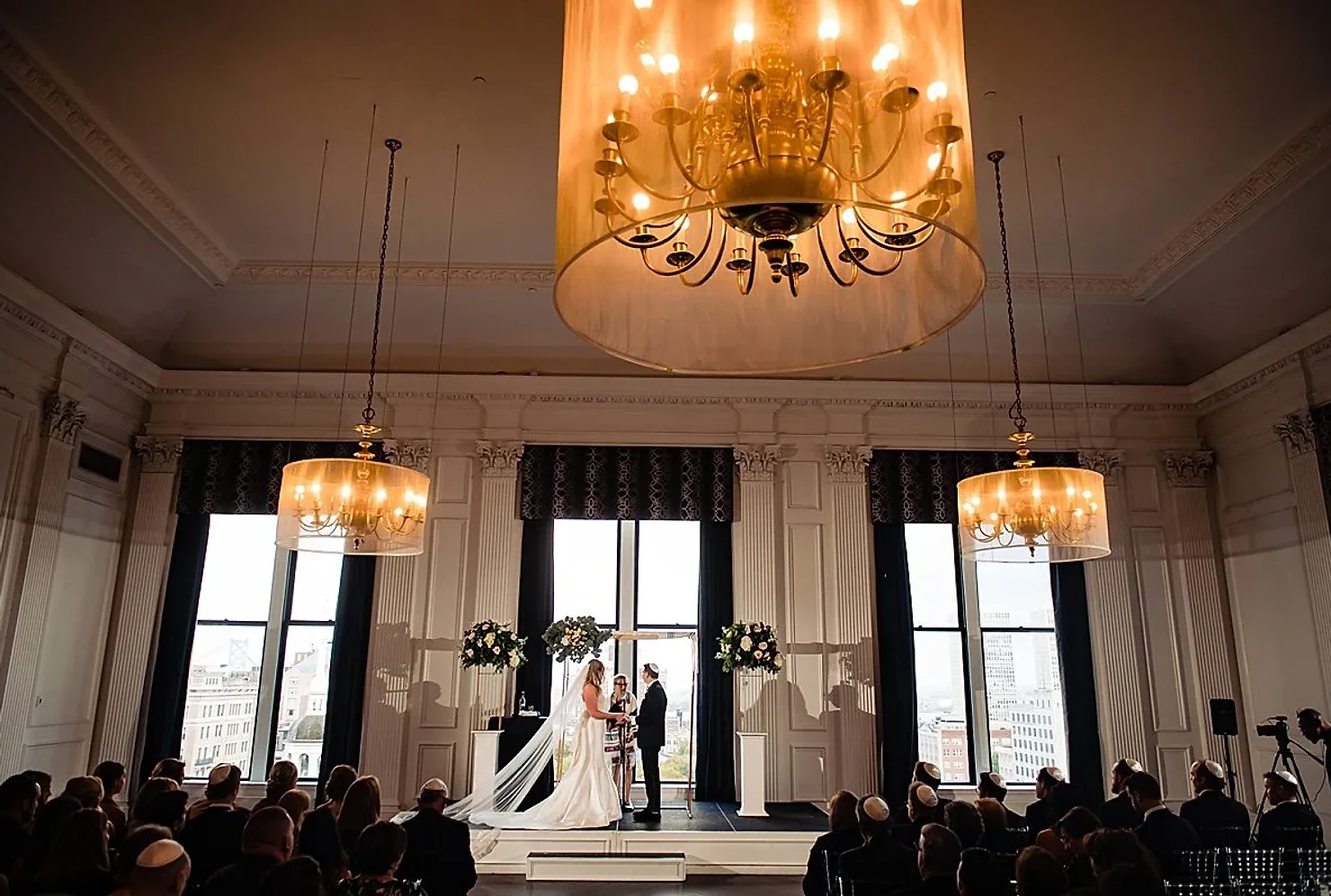 Philly wedding dj Bride and groom exchange vows in a grand hall with chandeliers, tall windows, and seated guests during a wedding ceremony.