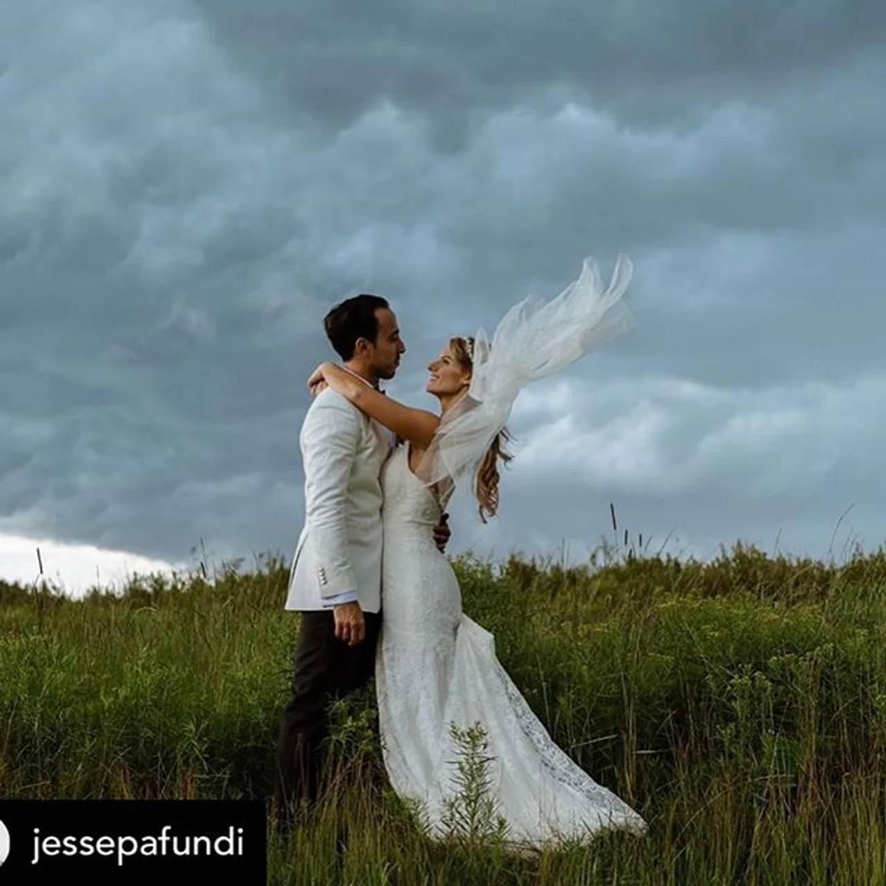 nyc catskills wedding dj Bride in white gown and groom in white jacket embrace in a grassy field under stormy clouds, veil blowing; jessepafundi