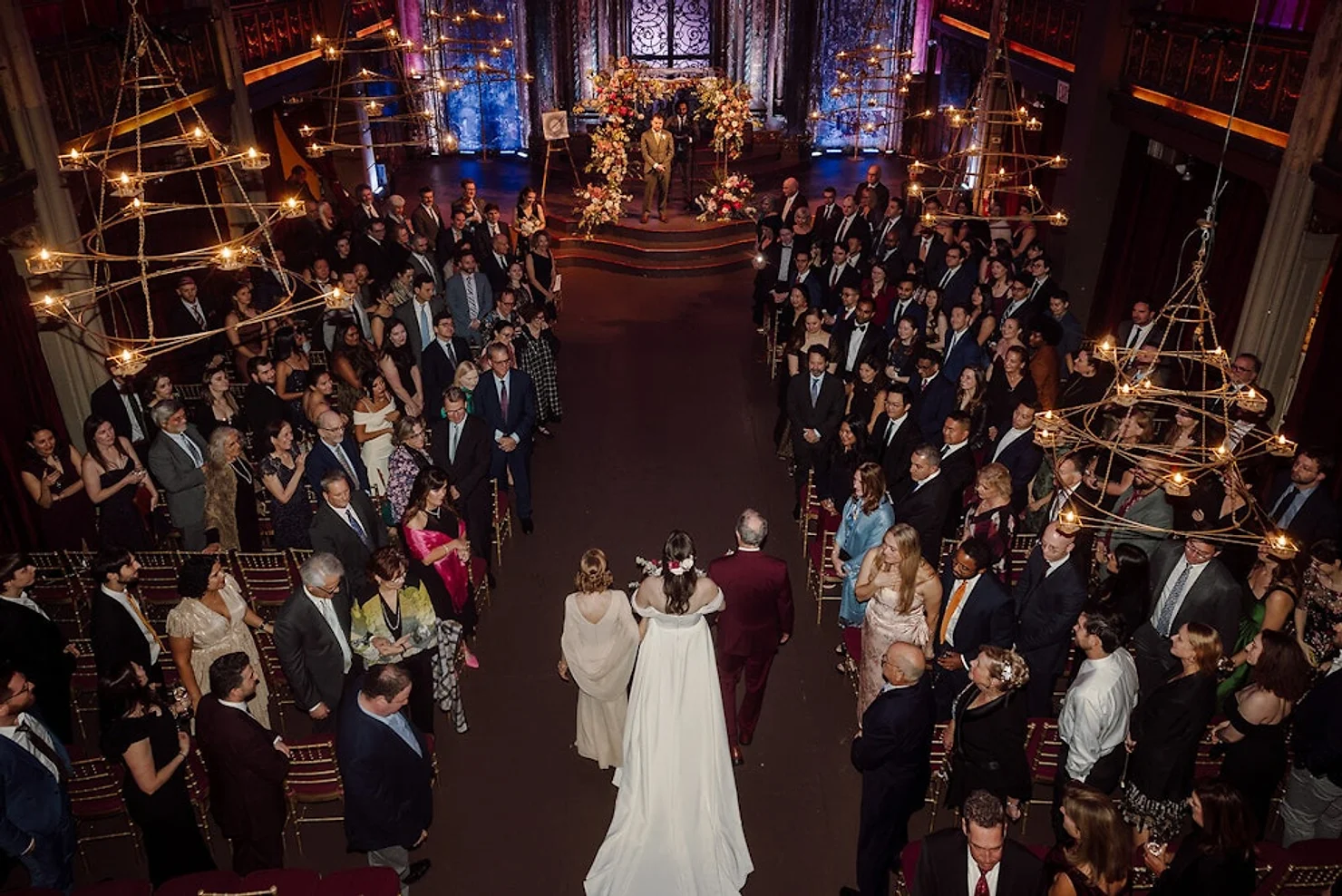 NYC wedding DJ Angel Orensanz Foundation Bride in white walks down a candlelit aisle with her parents at a formal wedding ceremony, guests standing in a dark ornate hall.