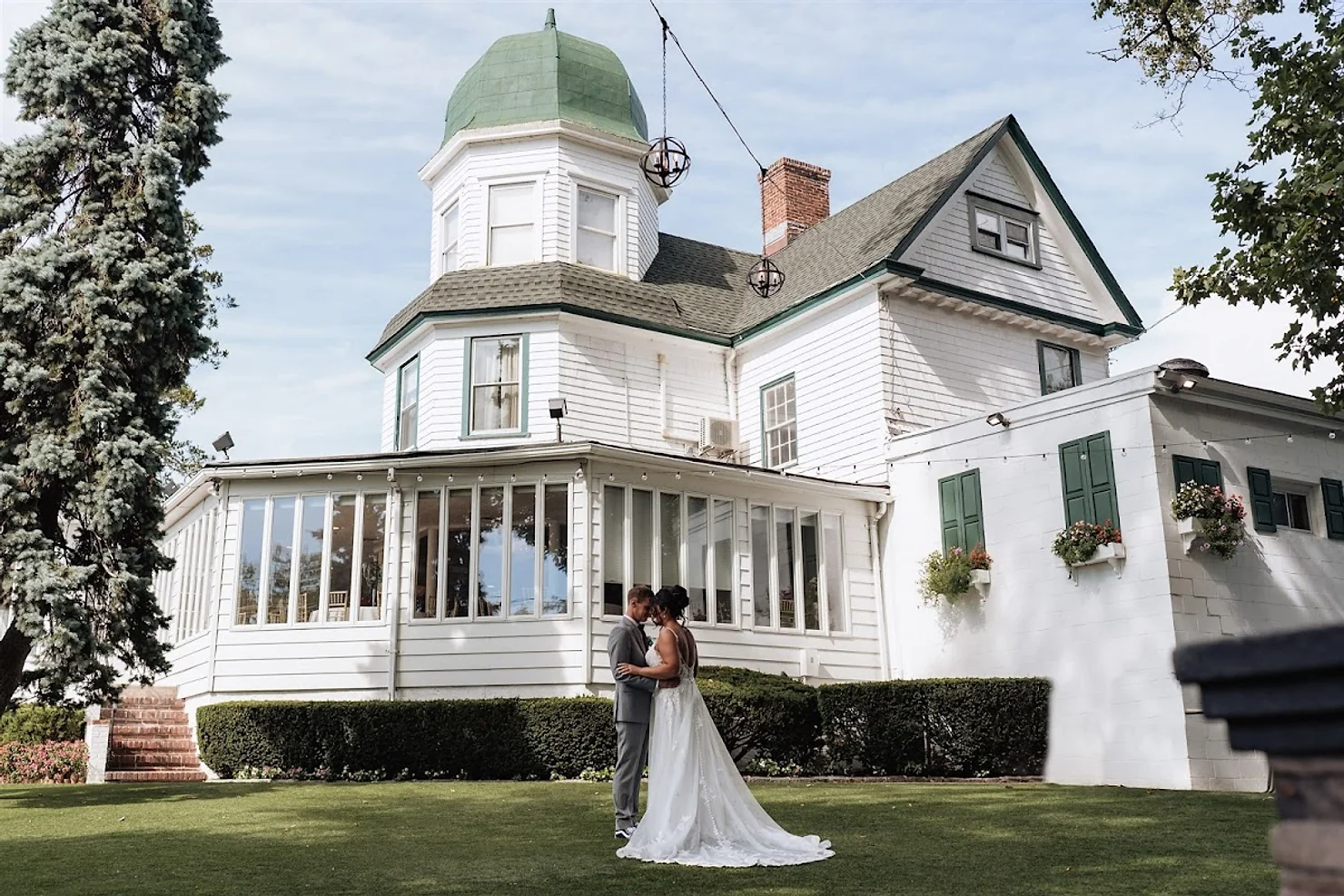 nassau county nyc wedding dj Bride and groom embrace on a lawn before a white Victorian house with a green dome, under a bright sky.