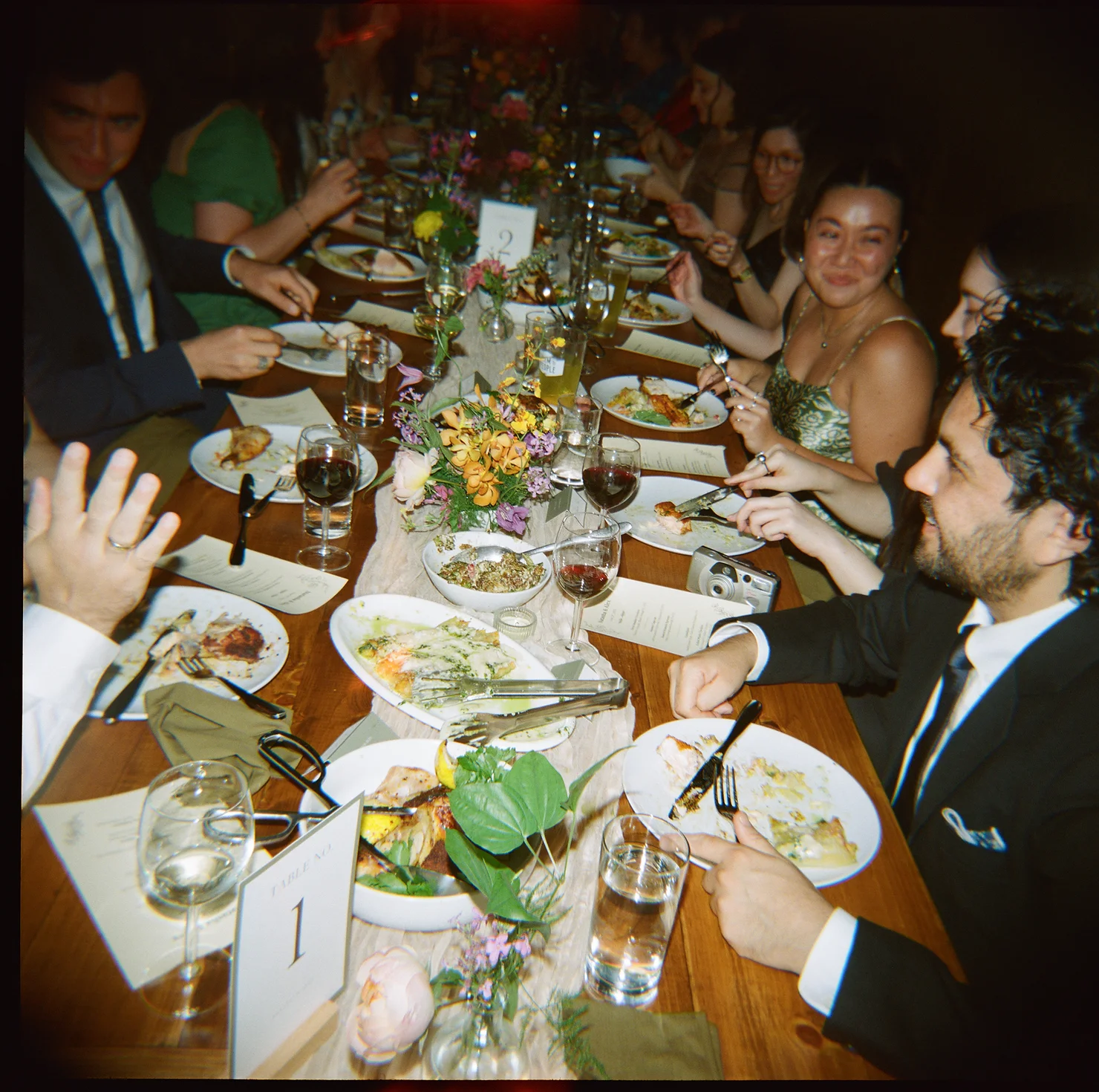 nyc non traditional wedding dj Friends in formal wear share dinner at a candlelit table with flowers, plates, wine glasses, and table numbers 1 and 2.