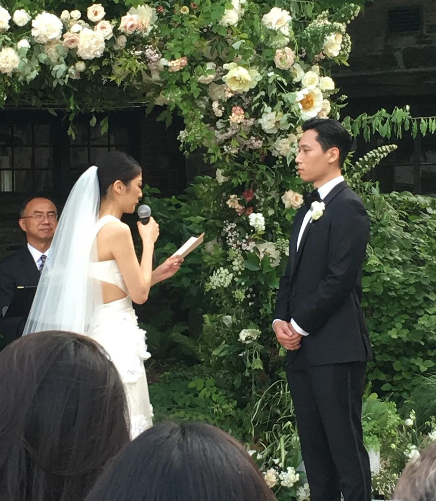nyc wedding dj Bride in white veil reads vows into microphone to groom in black tuxedo under a lush white-flower arch at an outdoor wedding.