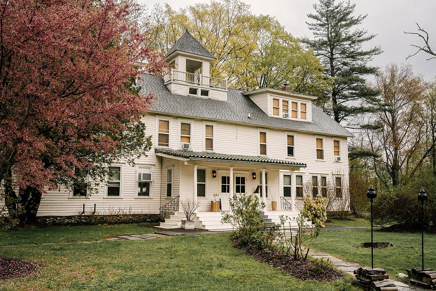 nyc ulster county wedding dj Large white house with gray roof and porch, framed by pink and green spring trees under an overcast sky.