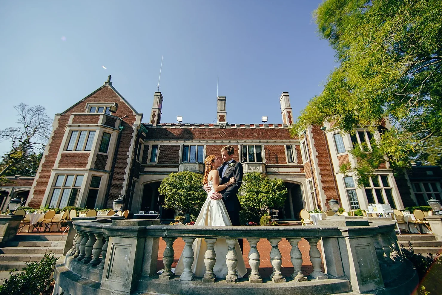 Waveny House Non Traditional Wedding DJ. Bride and groom embrace on a stone terrace outside a grand brick mansion under a clear blue sky, wedding mood romantic