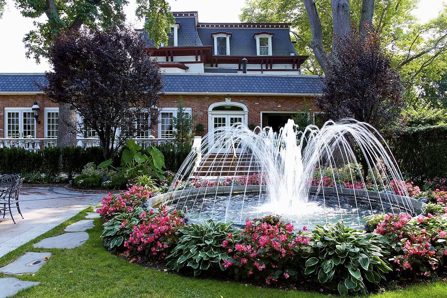 nassau county nyc wedding dj Brick mansion with slate roof behind a garden fountain, pink flowers, and trees in a bright landscaped courtyard.