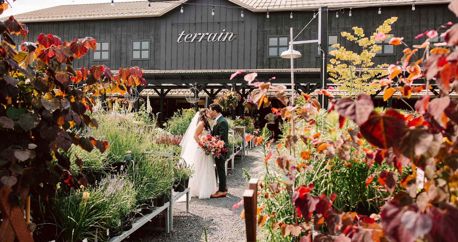 Bucks county wedding dj Bride and groom kiss in a lush garden outside a dark barn labeled terrain, framed by autumn leaves.