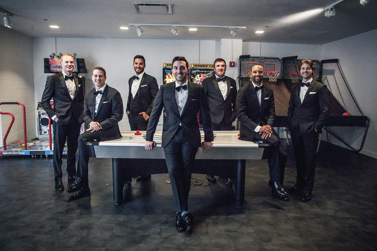 the loading dock non traditional wedding dj. Seven men in tuxedos pose around an air hockey table in a gray arcade game room, smiling under bright lights.