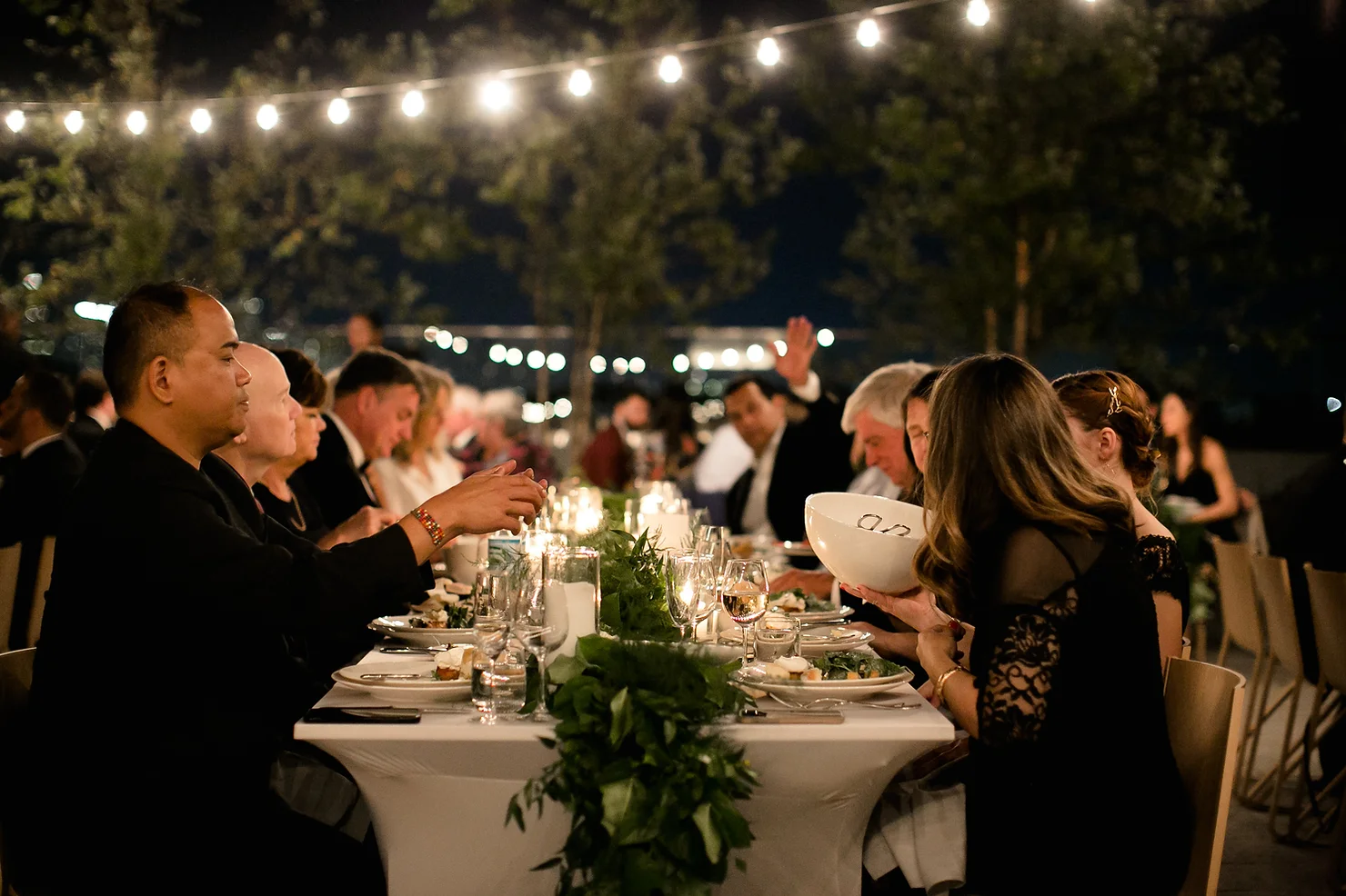 nyc wedding dj the william vale Guests dine at a candlelit outdoor dinner under string lights, sharing food at a long table in a warm evening mood.