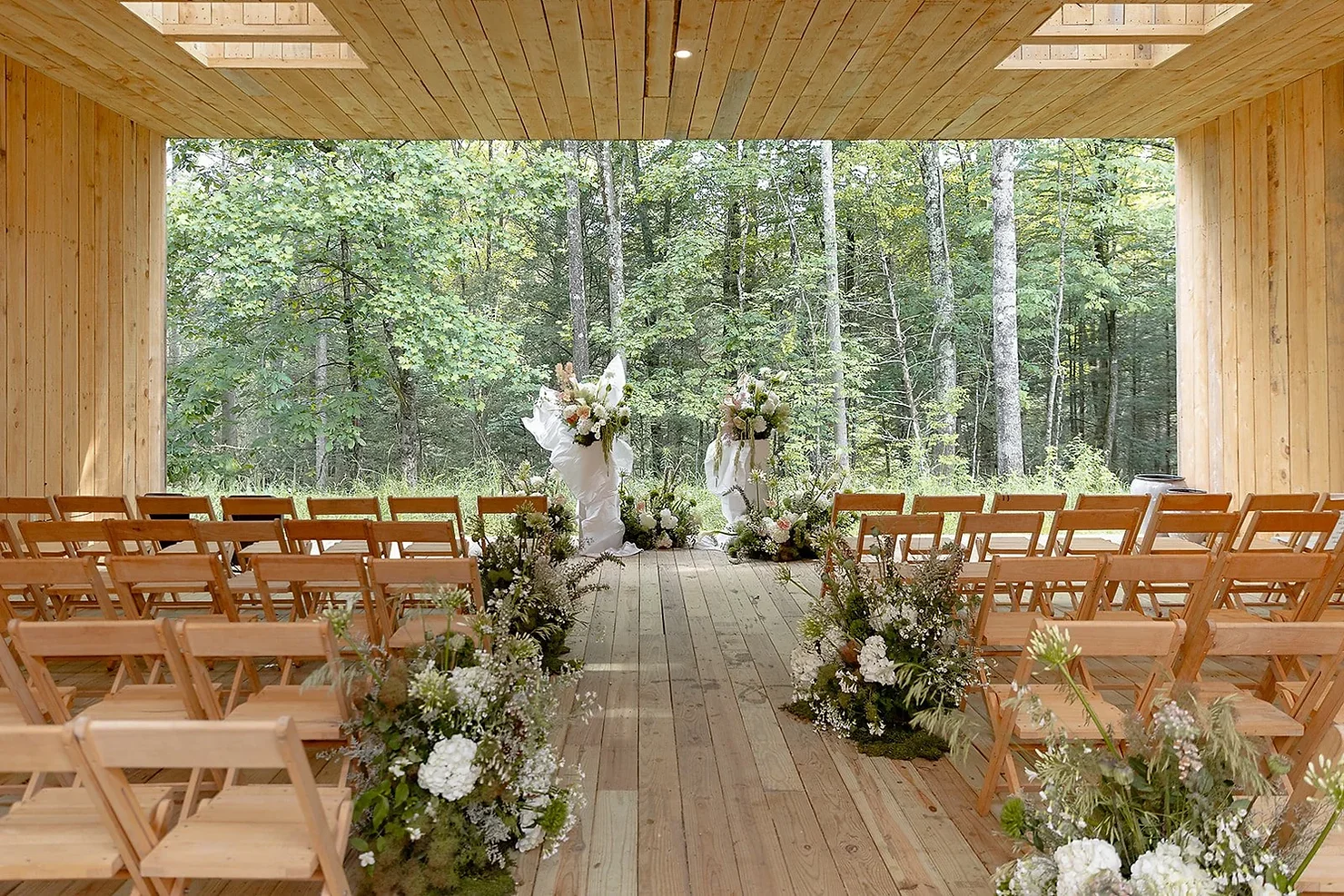 nyc wedding dj foxfire mountain house Empty rustic wedding ceremony space with wooden chairs and white floral arrangements overlooking a green forest outdoors
